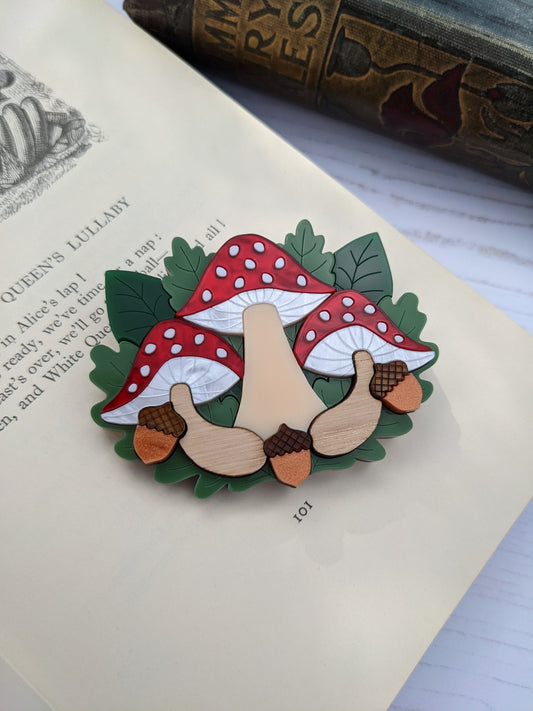 A laser cut acrylic brooch depicting stylized toadstools with red and white spots, green leaves, and brown caps, placed on a book page. Fly agaric