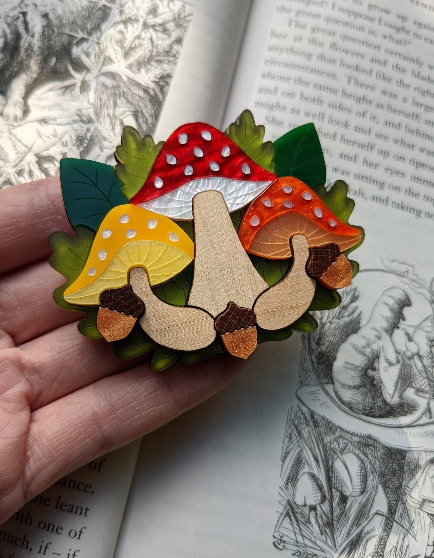 A colorful brooch shaped like toadstools with forest green, brown, and additional colors, held in a person's hand, with an illustration in the background.