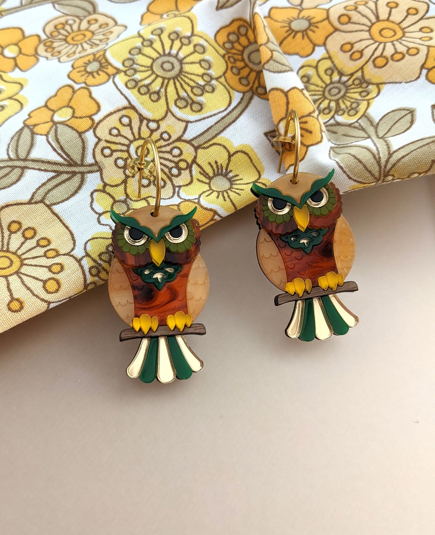 A pair of owl hoop earrings with a gold tone and multicolor design, displayed on a patterned fabric.