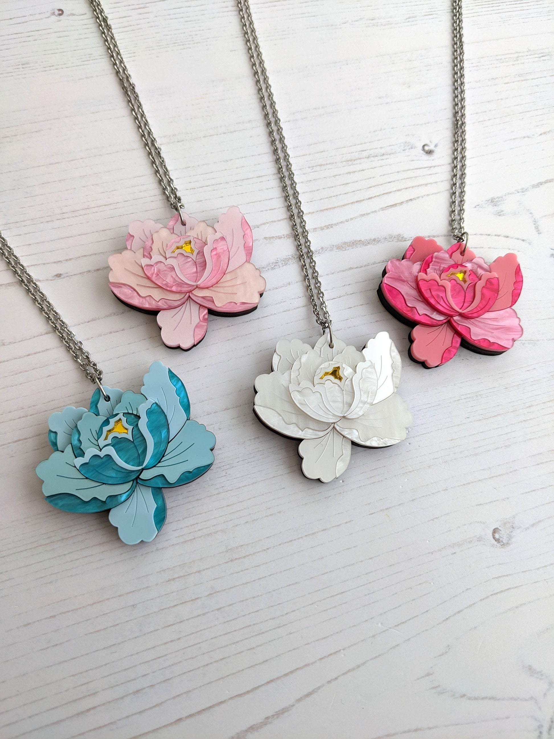 A collection of peony-shaped pendants in various colors (pink, blue, white, green) with a chain, displayed on a wooden surface.