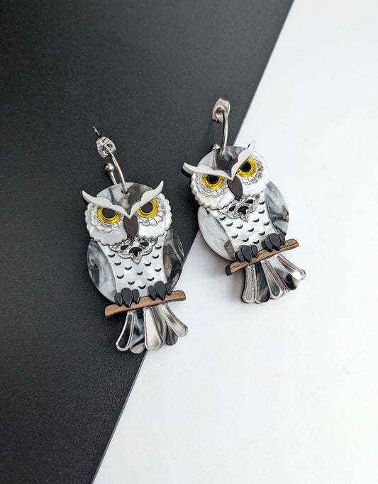 A pair of hoop owl earrings made with black and white acrylic, and wooden elements, displayed against a two-tone background.