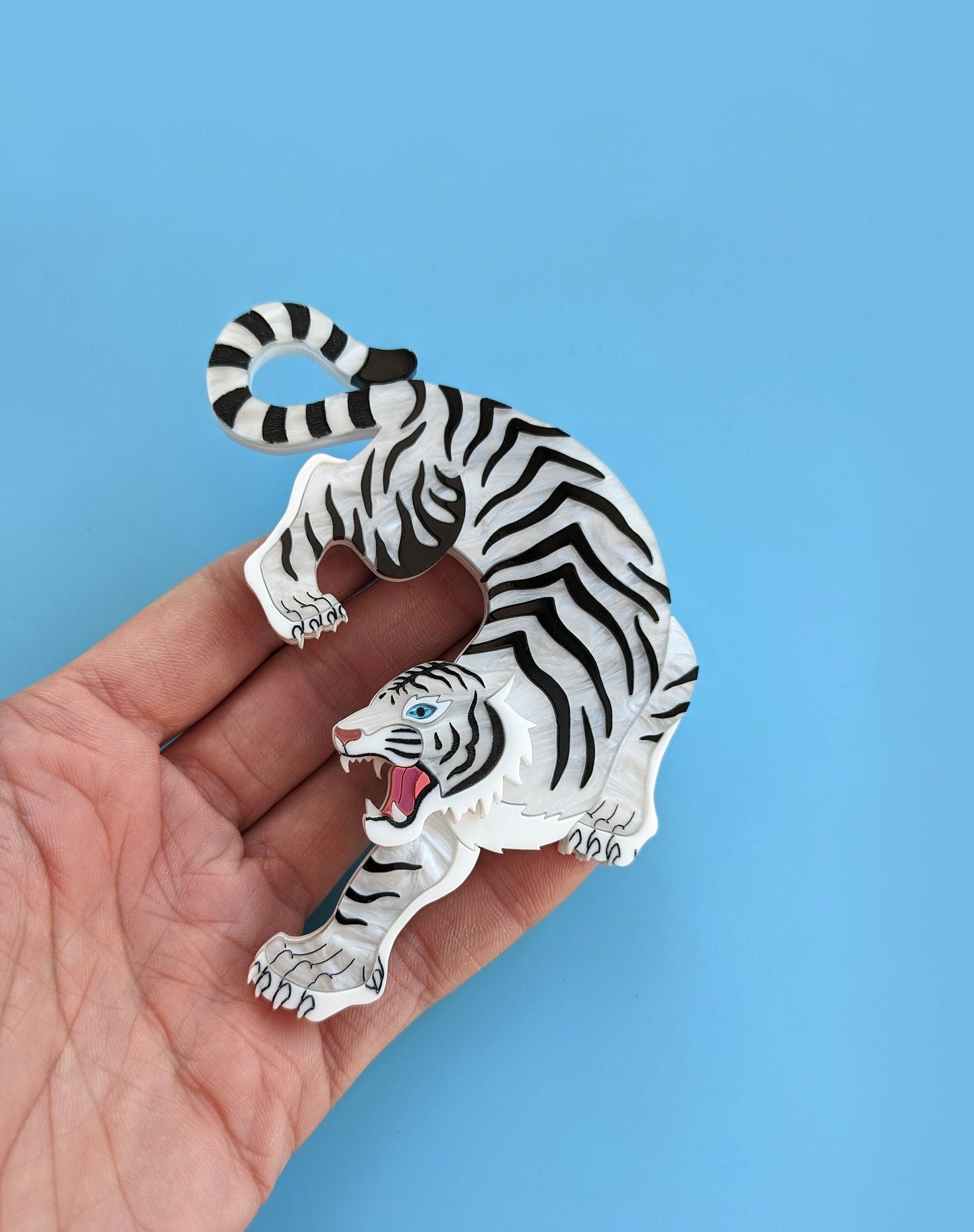 A hand holding a statement brooch designed in the shape of a white tiger with black stripes, featuring a blue mirror eye and metallic rose gold nose against a blue background.