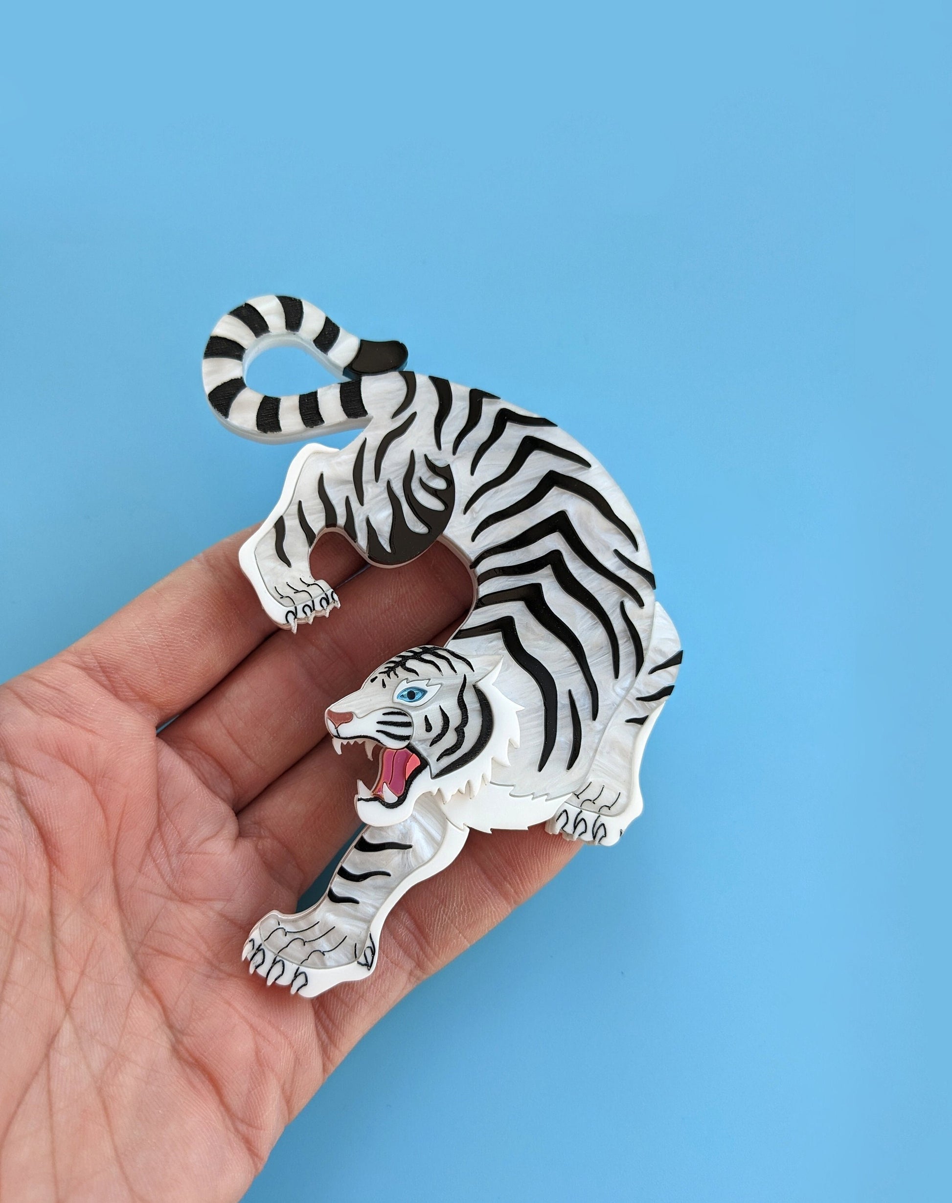 A hand holding a statement brooch designed in the shape of a white tiger with black stripes, featuring a blue mirror eye and metallic rose gold nose against a blue background.