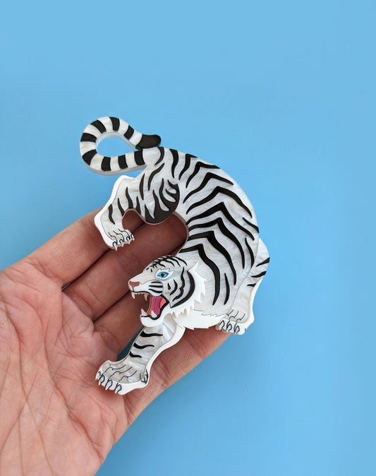 A hand holding a statement brooch designed in the shape of a white tiger with black stripes, featuring a blue mirror eye and metallic rose gold nose against a blue background.