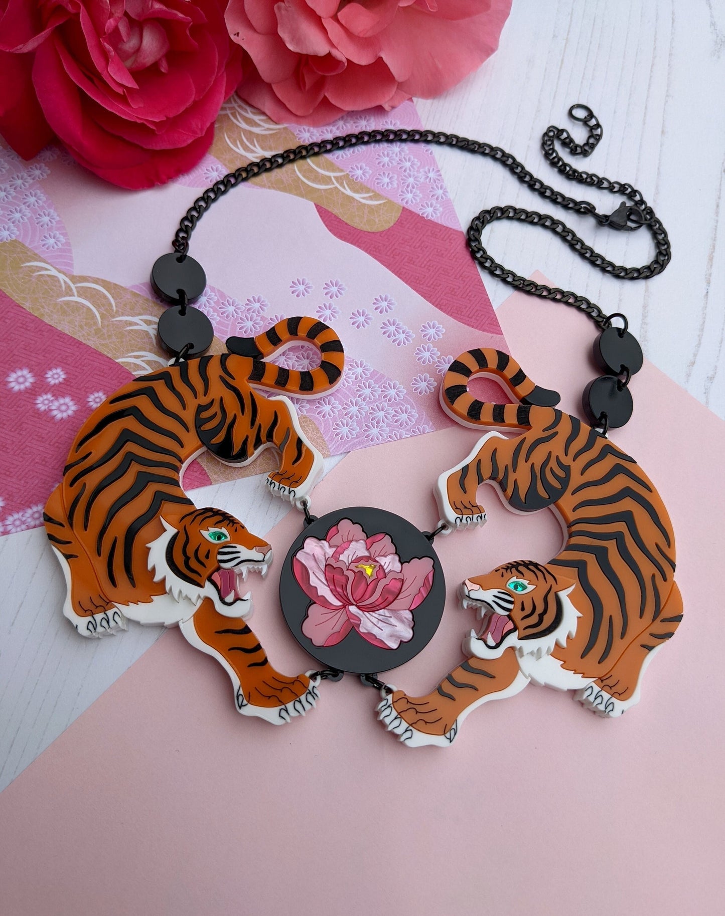 A striking orange and black acrylic necklace featuring two tiger designs with hand-inked details, and a central peony flower motif. The piece is displayed against a pink background with a floral pattern.