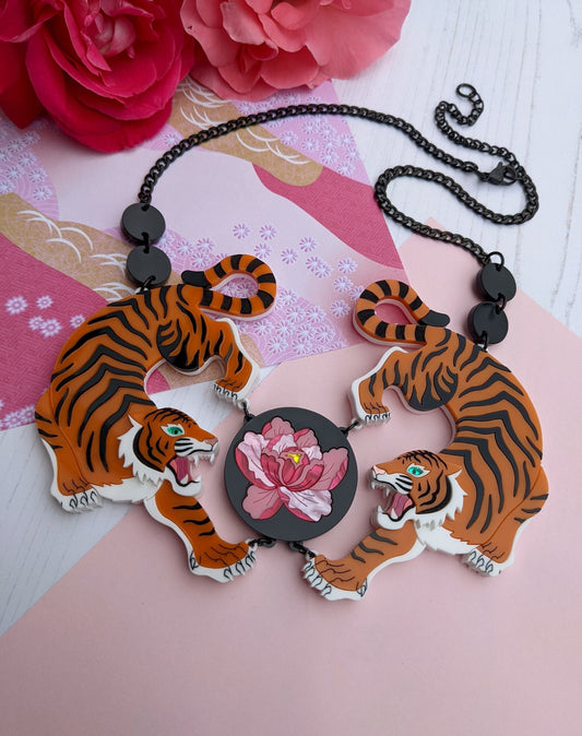A striking orange and black acrylic necklace featuring two tiger designs with hand-inked details, and a central peony flower motif. The piece is displayed against a pink background with a floral pattern.