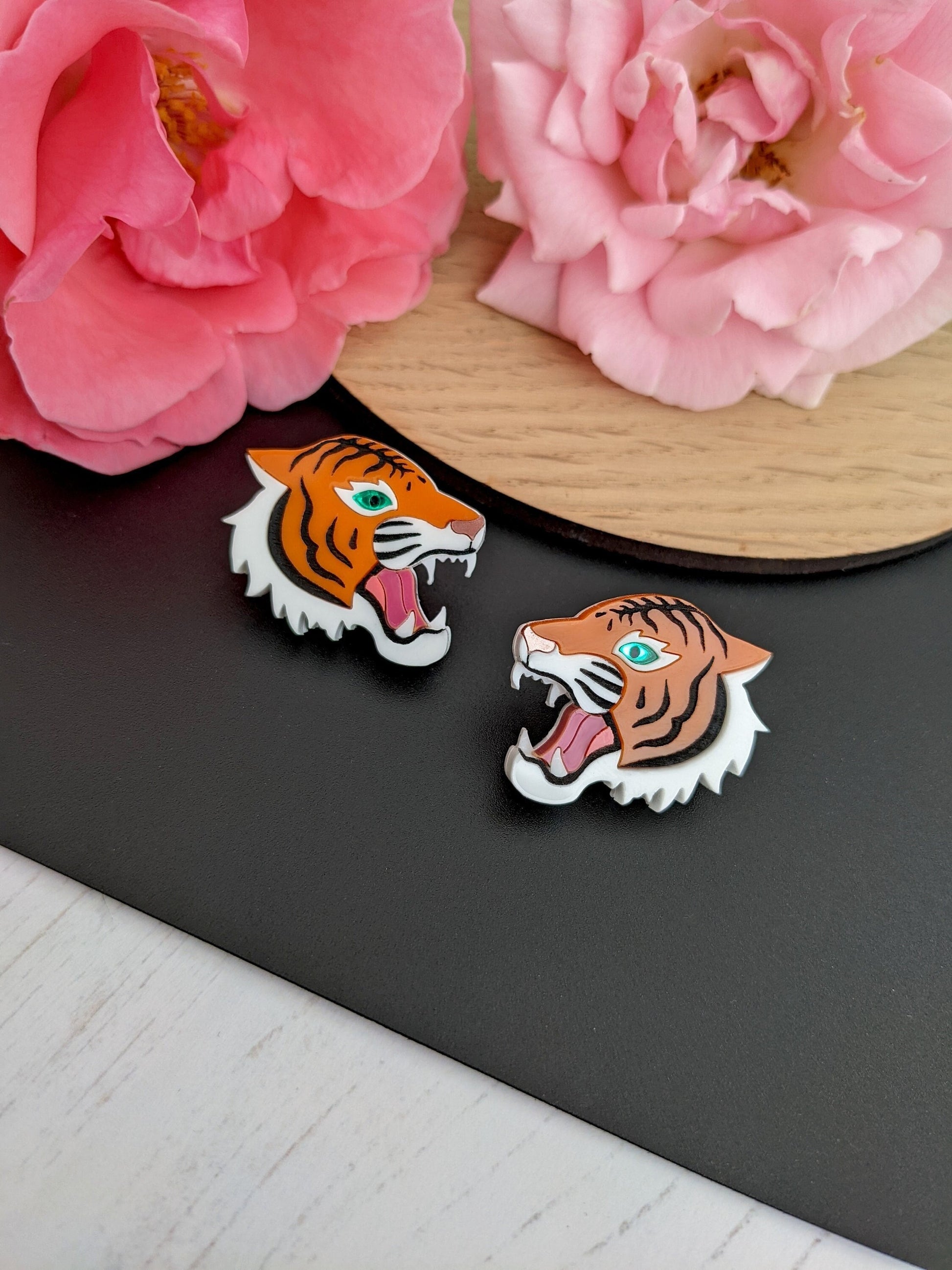A pair of orange tiger stud earrings with green eyes and rose gold metallic noses, displayed on a black surface with a wooden round and pink flowers in the background.