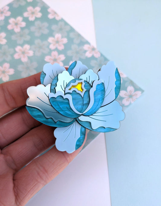 A hand holding a teal and blue acrylic brooch shaped like a peony flower, with a floral patterned background.