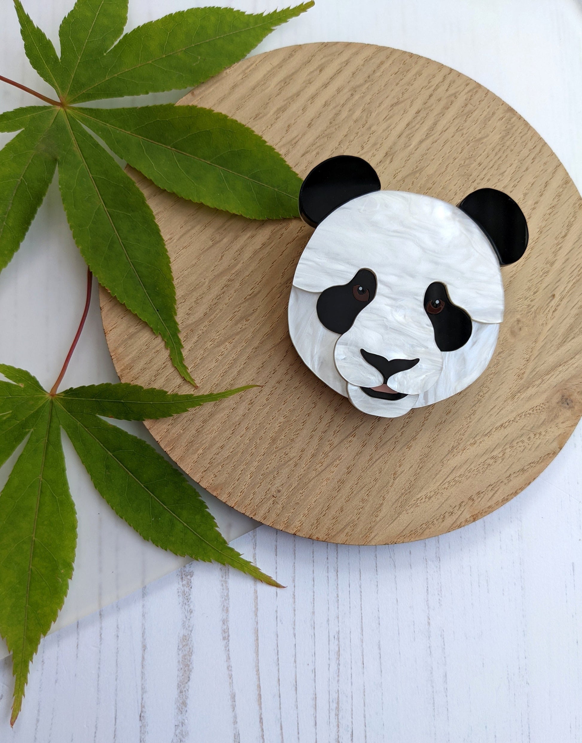 A brooch designed in the shape of a panda's head, made with plain and pearlescent marble acrylic, displayed on a wooden round coaster with a leaf to the side.