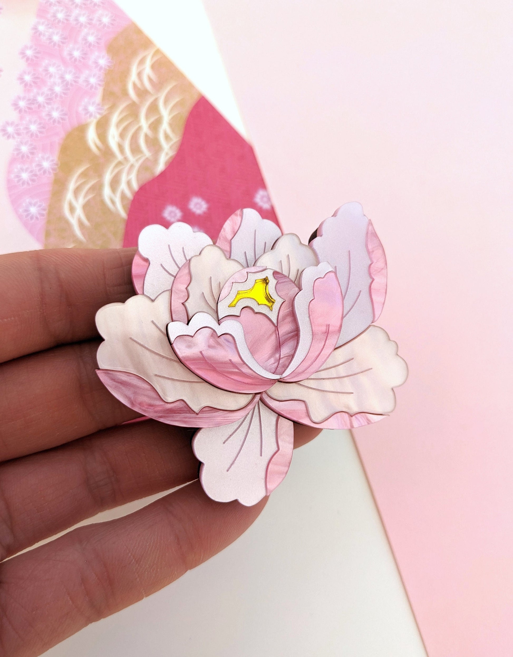 A delicate pink peony brooch with a floral design, held in a hand, showcasing its intricate detailing and pink tones against a light background.