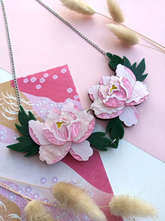 A delicate pink statement necklace featuring two stylized peony flowers, displayed on a flat surface with a stainless steel chain and green leaf charm.