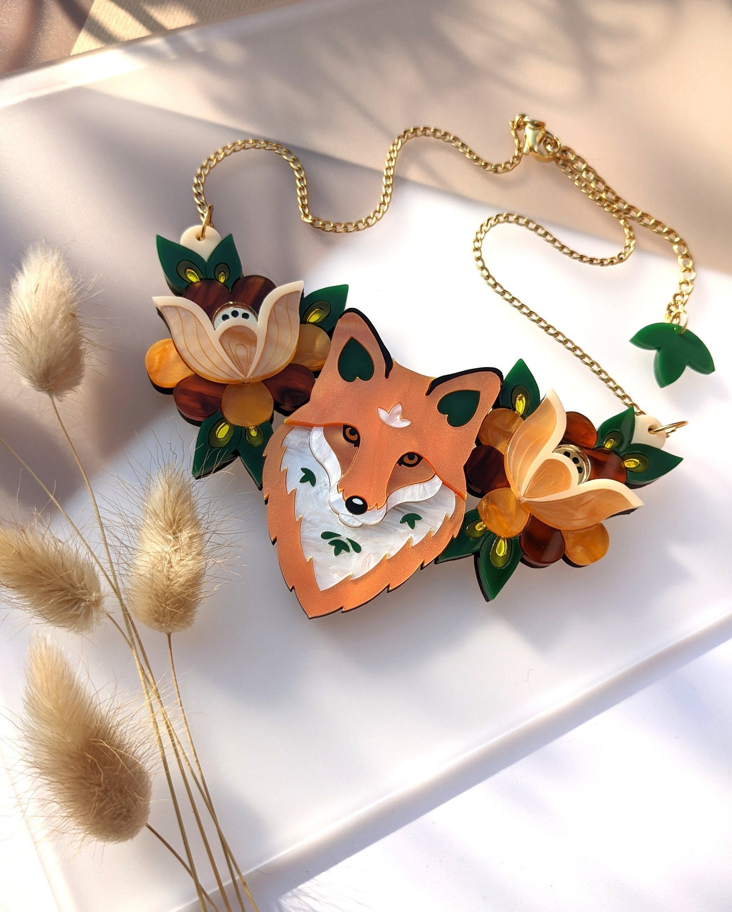 A bib necklace with a detailed fox design, surrounded by floral patterns, made from a combination of materials including pearl marble, tortoiseshell, and mirror acrylic, displayed on a white surface with dried plants beside it.