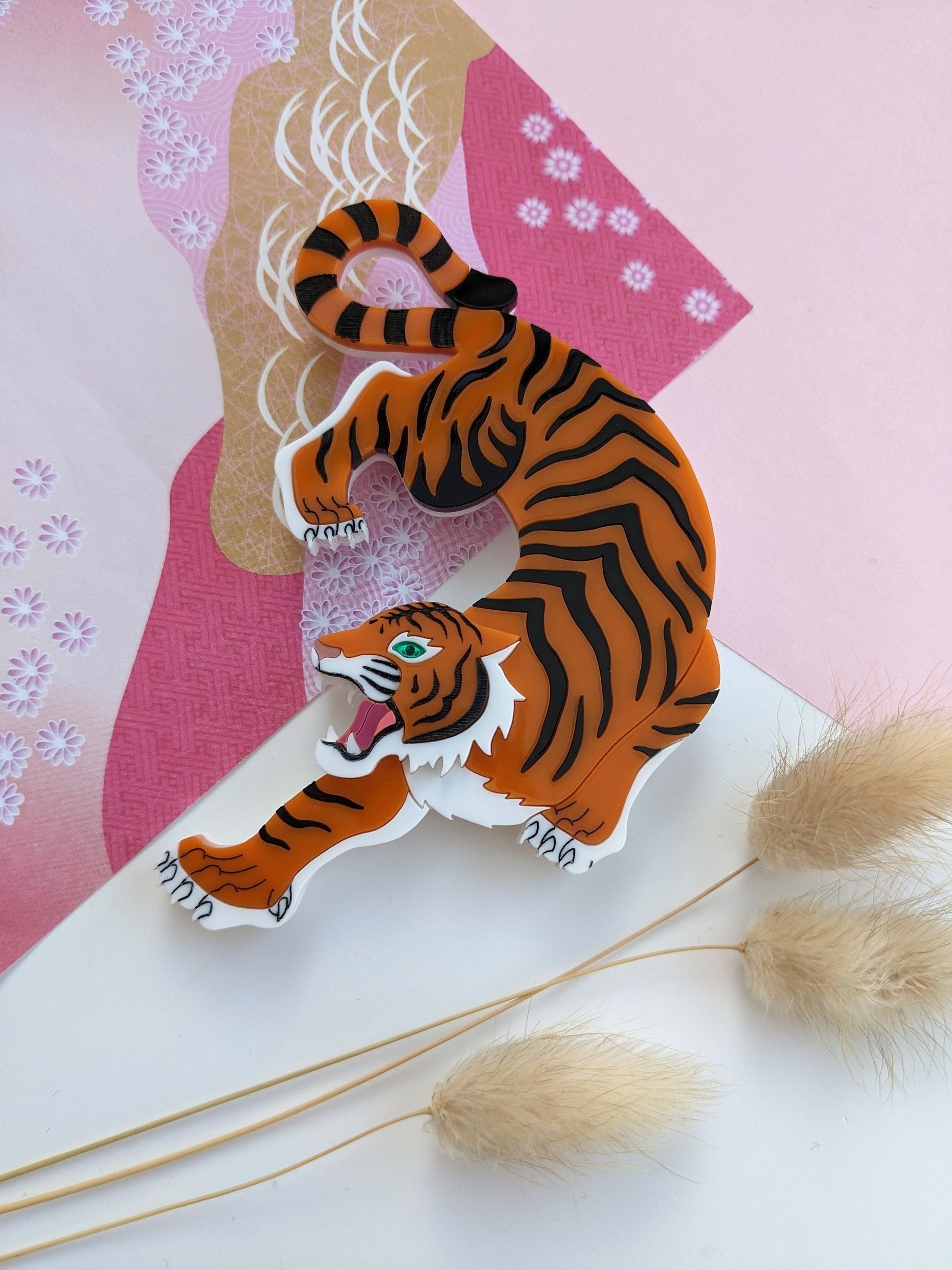 A large orange and black tiger brooch with a metallic rose gold nose and green mirror eye, displayed on a pink and white patterned background.