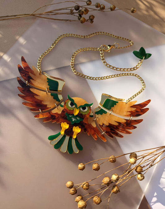 A vibrant, multicolored acrylic owl necklace with gold ion plated stainless steel chain, displayed on a white surface with decorative gold berries.