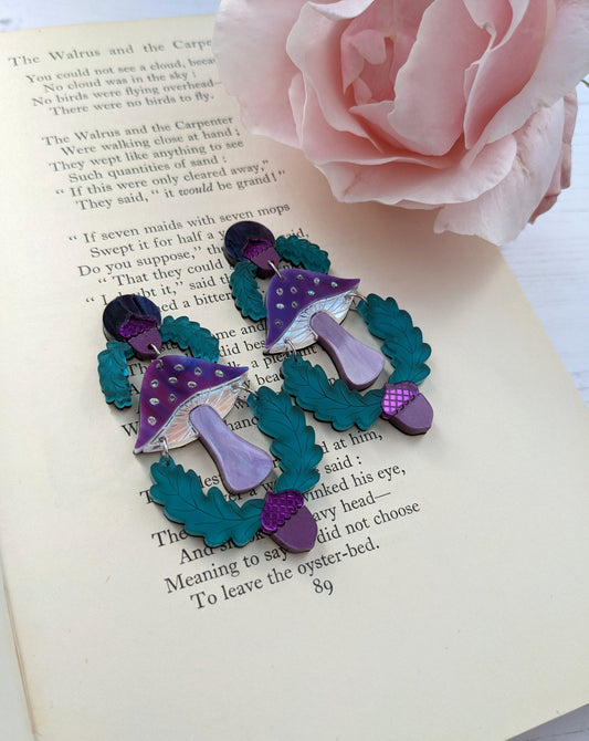 A pair of statement mushroom earrings, with purple and green caps, placed on an open book next to a pink rose.